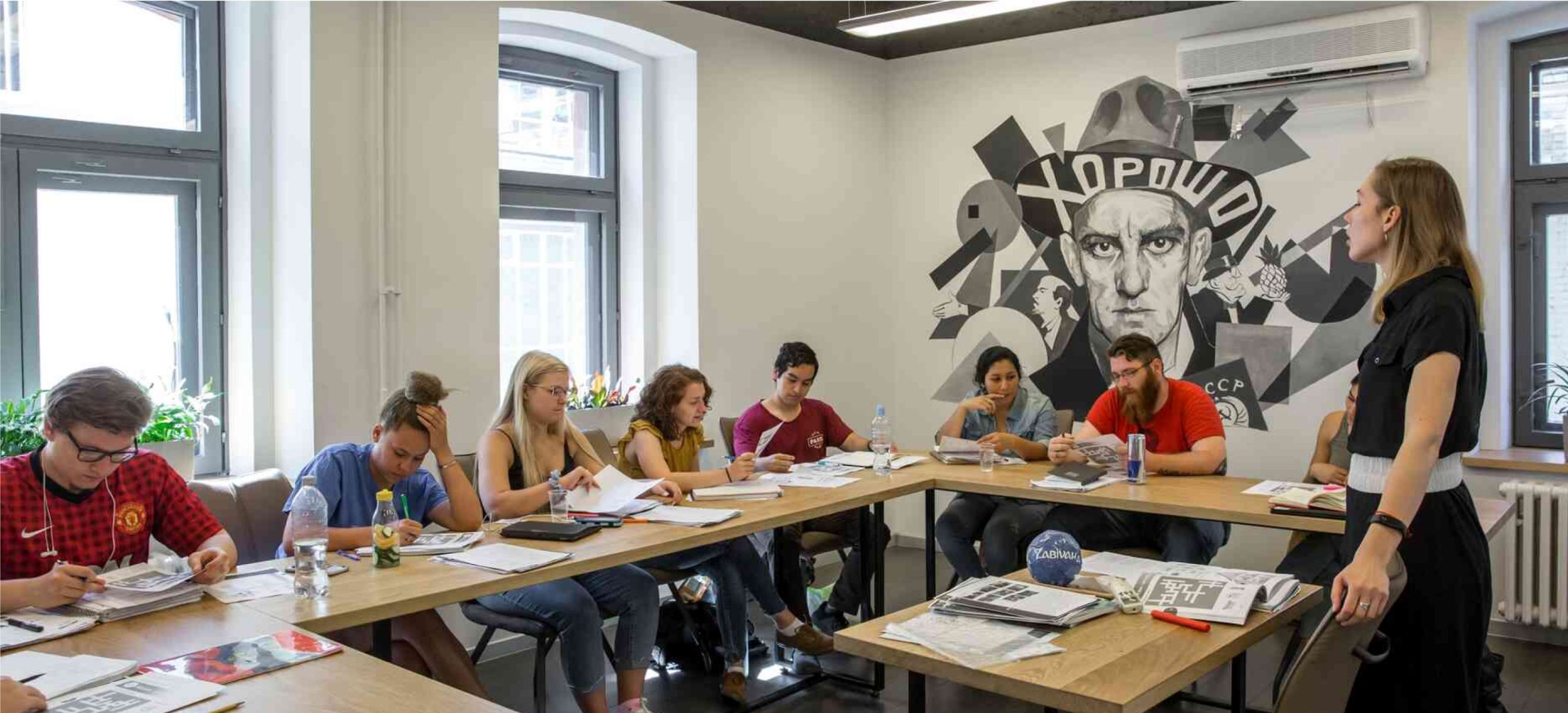 Teacher and students in a classroom with desks in a U-shape and a wall mural featuring the word ХОРОШО.