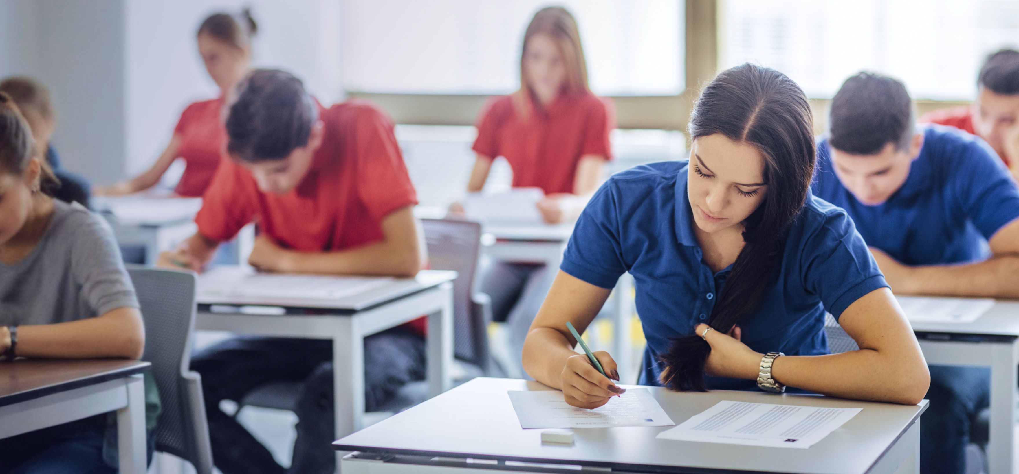 Students sitting at desks in a classroom taking an exam, with a woman in a blue shirt writing in foreground.