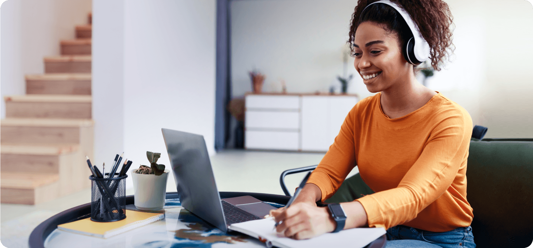 Smiling woman wearing white headphones sitting at a desk with a laptop and writing in a notebook.